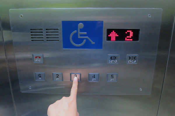 Pessoa apertando botão de elevador que possui texto no sistema Braille.