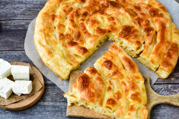 Foto de um banitsa, prato tradicional da Bulgária, fatiado e cubos de queijos sobre tábuas de madeira.
