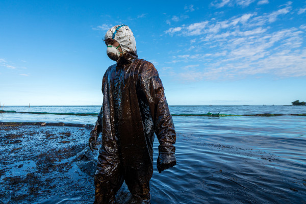 Foto de um homem, com roupas apropriadas e todo sujo, no oceano Índico, ajudando a limpar das águas uma mancha de petróleo.