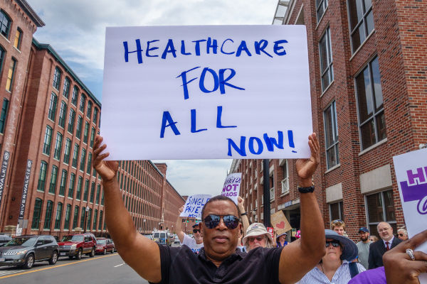 Homem segura cartaz onde se lê: “Healthcare for all now” (em tradução livre, “Saúde para todos agora”).