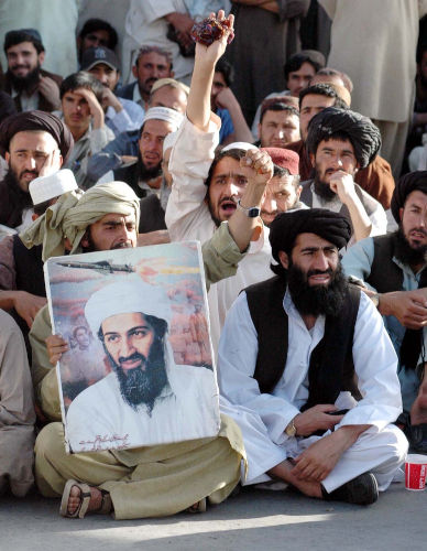 Homens muçulmanos reunidos e sentados; um deles segura o retrato de Osama bin Laden.