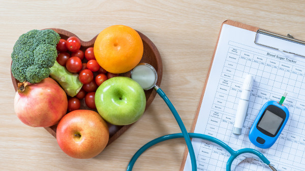 Frutas e verduras sobre um prato de coração próximo a um glicosímetro sobre uma prancheta.