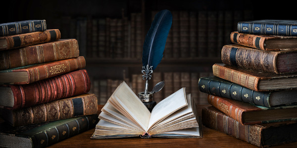 Um livro em branco aberto em frente a uma caneta-tinteiro de pena e no meio de duas pilhas de livros antigos.