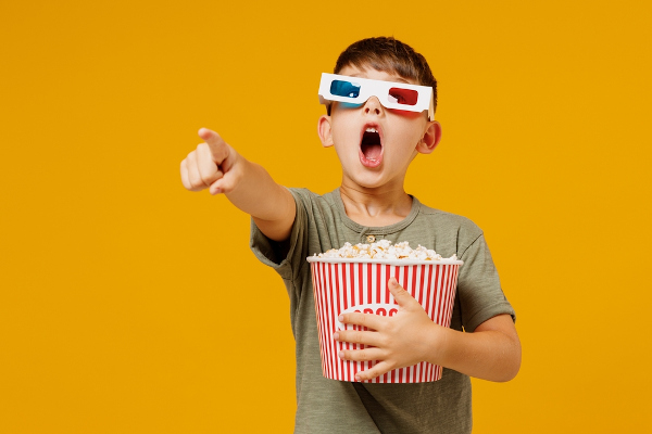Criança com óculos 3D segurando um balde de pipoca e apontando para algum lugar com expressão de impressionamento.
