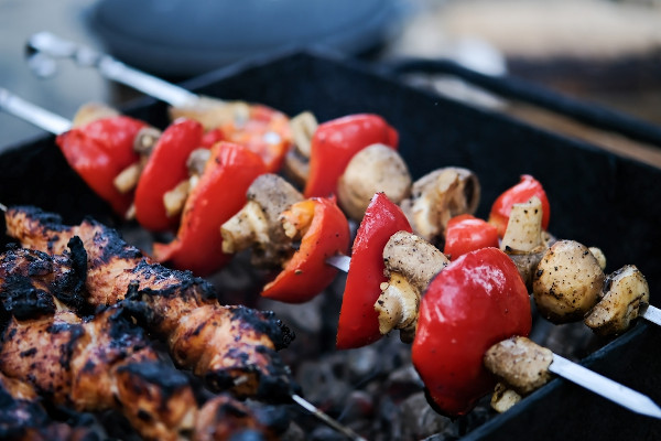 Vista aproximada de carne, cogumelo e pimentão sendo assados em espetos em uma churrasqueira com carvão vegetal.