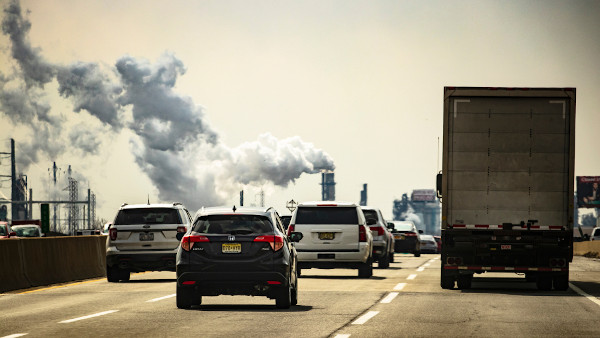 Região urbana com a presença de um caminhão, de vários carros e de indústrias lançando fumaça na atmosfera.