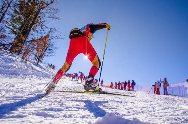 Atleta em competição de esqui nórdico com roupa vermelha e torcedores ao fundo. 