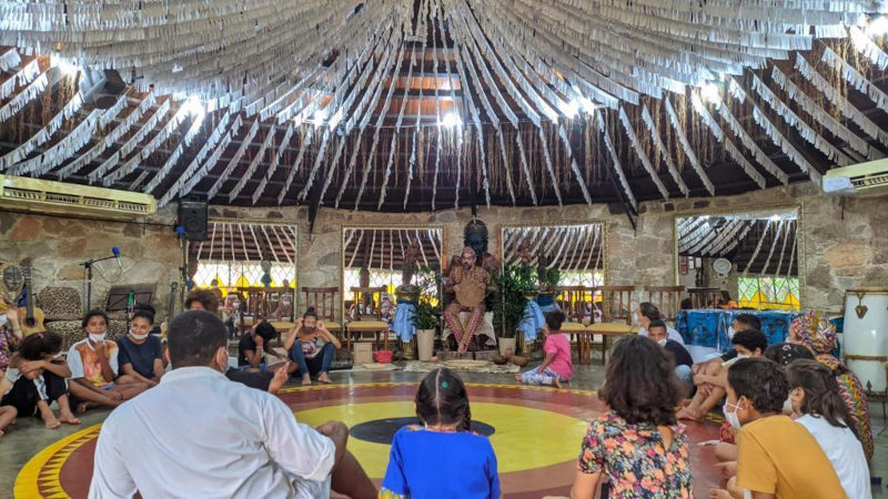 Espaço redondo com crianças sentadas descalças com mácara. Há um palco com pessoas adultas se apresentado musicalmente