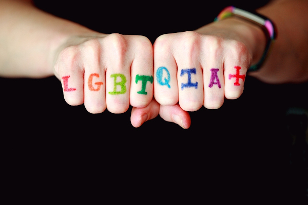Letras que compõem a sigla LGBTQIA+ pintadas em dedos das mãos.