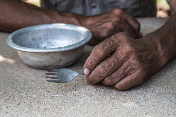 Pessoa sentada segurando um garfo próximo a um prato de alumínio vazio; a pobreza é uma causa da desnutrição.