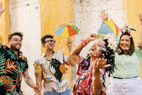 Pessoas sorrindo, jogando confetes e segurando guarda-chuvas coloridos no Carnaval, festa que deu origem às marchinhas.