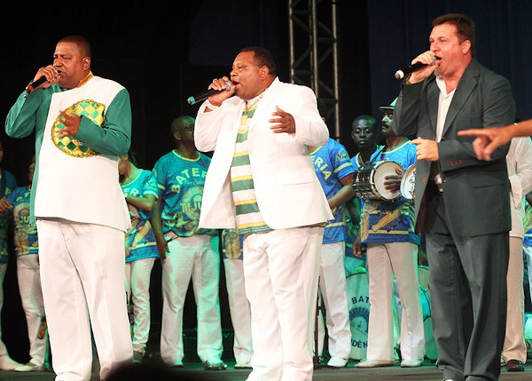 Três homens segurando microfones e cantando o samba-enredo de uma escola de samba.