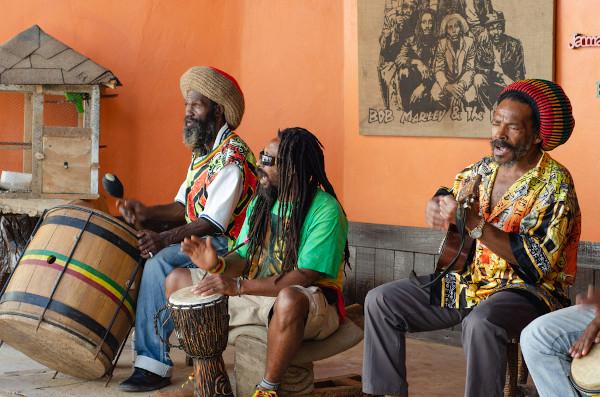 Três homens negros sentados tocando instrumentos de origem africana em tributo a Bob Marley, rei do reggae.