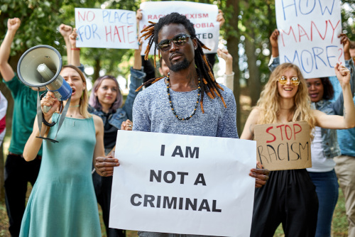 Grupo de pessoas realiza protesto. Homem ao centro segura cartaz, onde se lê: “I am not a criminal”.
