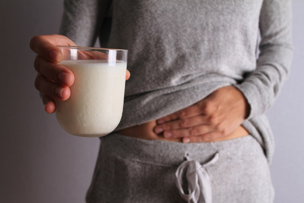 Mulher com uma mão sobre a barriga e, com a outra, segurando um copo com leite, alimento que pode causar gases (flatulência).