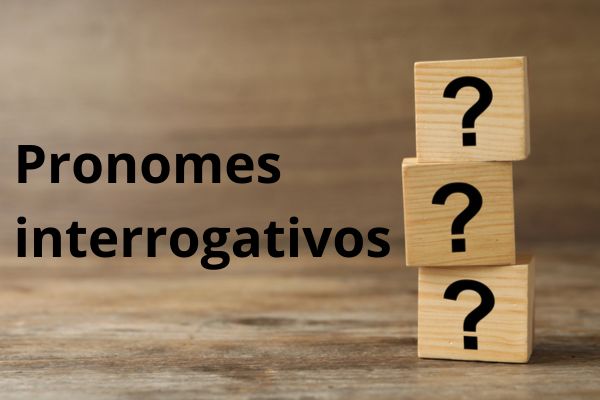 Três dados de madeira com pontos de interrogação empilhados ao lado do texto “pronomes interrogativos”.