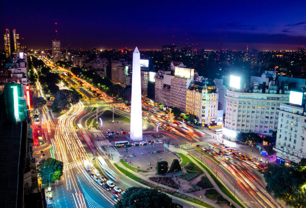 Vista aérea noturna do Obelisco de Buenos Aires no centro de uma avenida movimentada da metrópole argentina. 