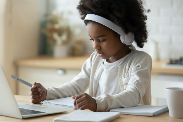 Estudante criança negra escrevendo com fone de ouvido em frente a um notebook branco