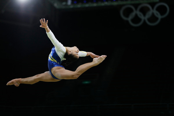 Flávia Saraiva, mulher branca, em salto de ginástica artística com abertura de quadril e flexão para trás do tronco.