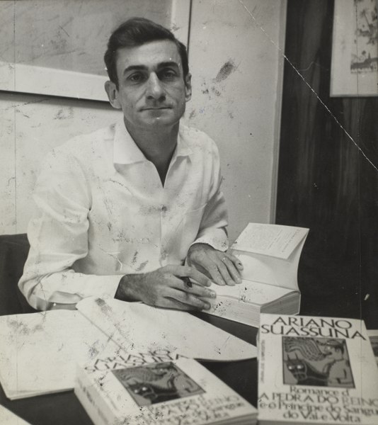 Fotografia em preto e branco de Ariano Suassuna autografando alguns livros.