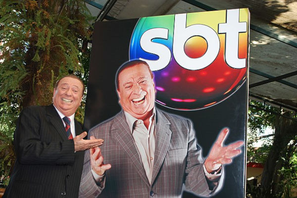 Raul Gil sorrindo ao lado de uma imagem com sua foto e a logomarca do SBT.