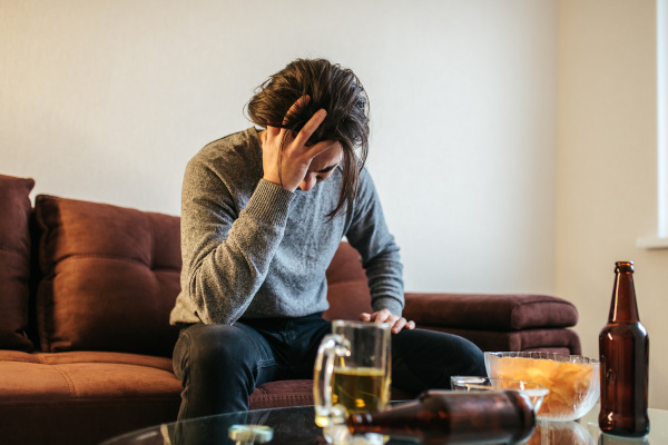 Homem coloca mão na cabeça diante de várias bebidas, em referência ao alcoolismo.  