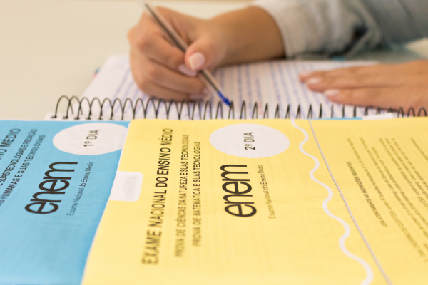 Mãos escrevendo em um caderno ao lado de provas do Enem, em texto sobre redação pronta. Mãos escrevendo em um caderno ao lado de provas do Enem, em texto sobre redação pronta.