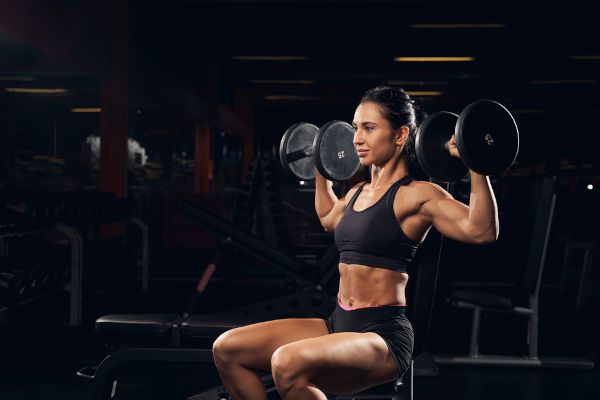Mulher faz desenvolvimento com halteres, exerc&iacute;cio de muscula&ccedil;&atilde;o para ombros. [imagem_principal]