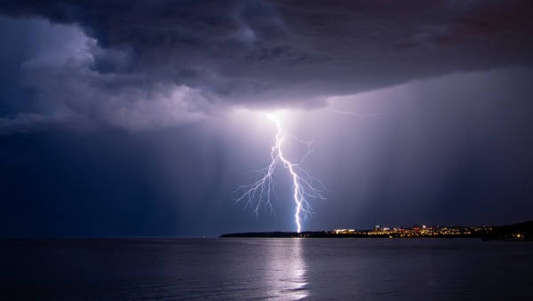 Raio ilumina céu noturno com muitas nuvens, acima de um lago e com luzes de cidade ao fundo. 