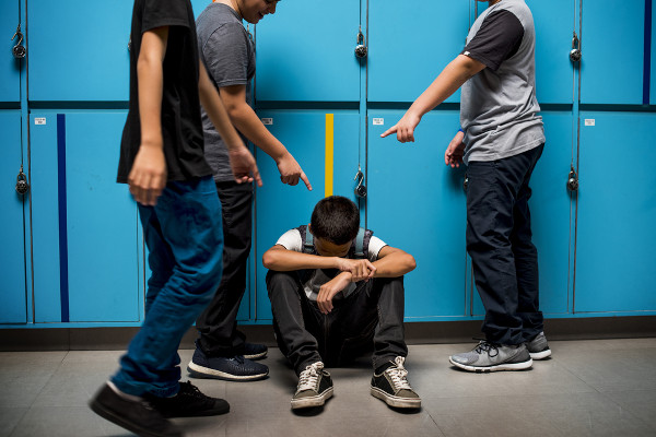 Três garotos de pé apontando para um garoto sentado e cabisbaixo, em referência ao bullying.
